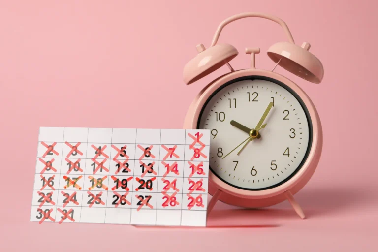 A pink alarm clock sits next to a calendar with many dates crossed out in red, symbolizing missed periods and the disruption of time associated with amenorrhea.