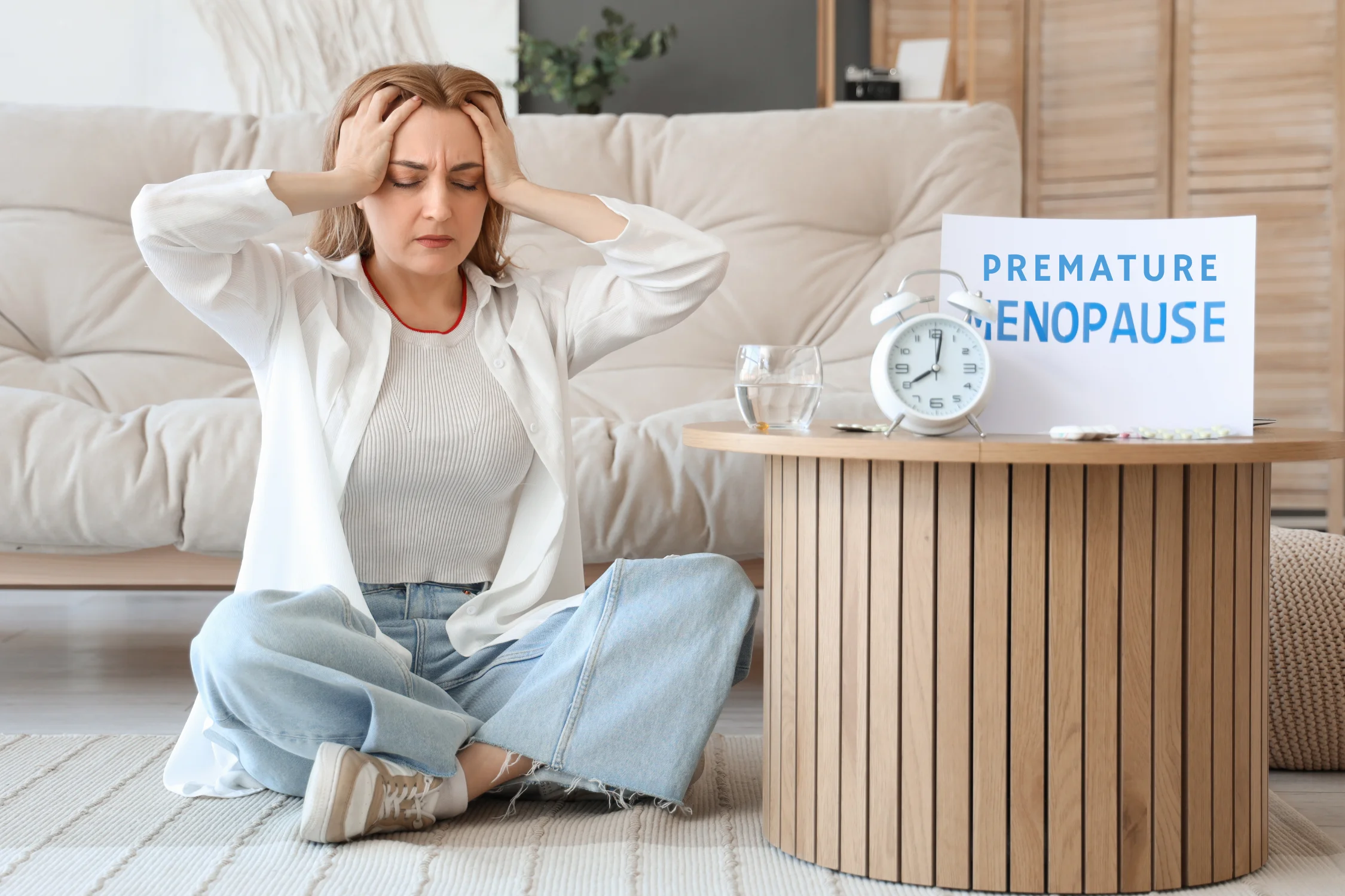 Middle-aged woman holding her head in discomfort while sitting next to a table labeled "Premature Menopause," symbolizing the emotional and physical symptoms of early menopause.
